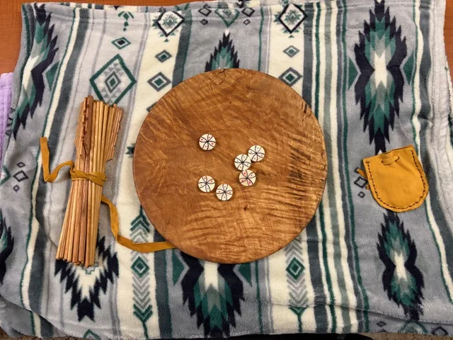 A wooden bowl with dice pieces, counting sticks, and a small leather pouch arranged for the Mi’kmaq dice and bowl game during the Wellness Gathering.